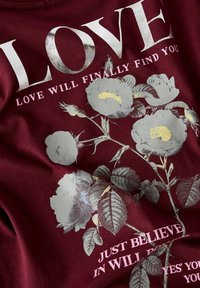 Camiseta burdeos con texto en plata y rosa; presenta un diseño floral con rosas grises y hojas verdes; textura de tela suave.