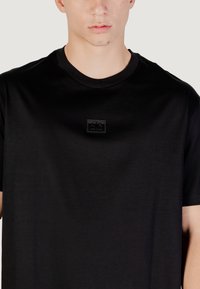 T-shirt in cotone nero con scollatura rotonda e maniche corte. Presenta un piccolo patch logo rettangolare nero sul petto. Texture liscia.