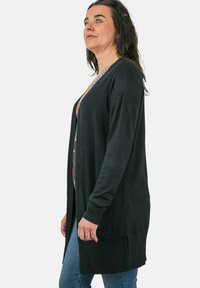 Cárdigan negro, diseño largo con textura acanalada, de apertura frontal, con dos bolsillos, usado sobre una camisa con estampado, combinado con jeans azules.
