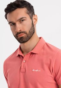 Korálová polo košile z bavlny, s límečkem a krátkými rukávy. Výrazný detail: bílé vyšívané logo na hrudi. Hladká textura.