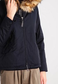 Tummansininen puffatakki, jossa on tekoturkisvuori hupussa, vetoketjusuljenta, sivutaskut ja sileä pinta. Ominaisuuksiin kuuluu tyylikäs muotoilu ja hieman lyhennetty malli.