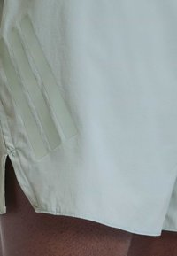 Αθλητικά σορτς ανοιχτού χρώματος με τρεις παράλληλες αντανακλαστικές ρίγες και μικρή πλάγια σχισμή, φορεμένα πάνω σε δέρμα με ορατή τρίχα.