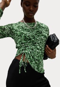 Groene gebloemde lange mouwen top met gerimpelde details, gecombineerd met een zwarte clutch. De stof heeft een gestructureerd, bloemmotief en zijstrikjes.