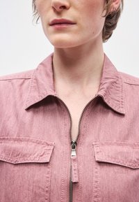 Rózsaszín farmerdzseki elülső cipzárral, gallérral és két mellzsebbel. Az anyag puha tapintású és jól látható varrási részletekkel rendelkezik.