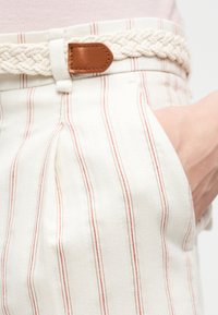 Λευκό παντελόνι με λεπτές κόκκινες κάθετες ρίγες, φορεμένο με πλεκτή μπεζ ζώνη που διαθέτει καφέ δερμάτινη αγκράφα, χέρι στην τσέπη.
