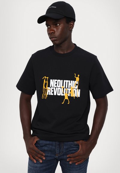 Sort bomulds T-shirt med hvid og gul grafisk tekst "NEOLITHIC REVOLUTION" med stiliserede figurer. Parret med blå denim jeans.
