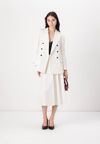 White double-breasted blazer with black buttons, paired with a matching midi skirt and a black top. Model holds a small burgundy handbag.