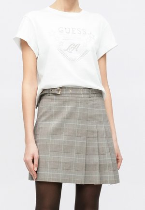 Woman wearing a white short-sleeve shirt with "GUESS LA" text and a gray plaid skirt with a belt, standing against a plain background.