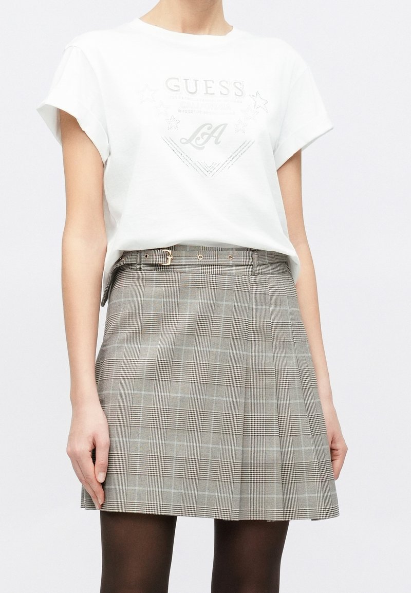 Femme portant une chemise blanche à manches courtes avec le texte "GUESS LA" et une jupe grise à carreaux avec une ceinture, debout devant un fond uni.