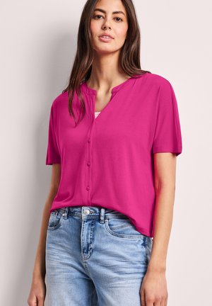 Kvinde iført en knaldrød kortærmet knapbluse, stoppet ned i lyseblå denimjeans, stående foran en ensfarvet lys baggrund.