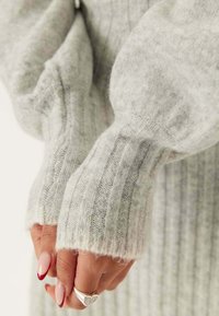 Maglione lavorato a maglia grigio chiaro con texture a coste e vestibilità comoda. Presenta maniche lunghe e larghe e una finitura leggermente pelosa. Mani con unghie curate visibili.