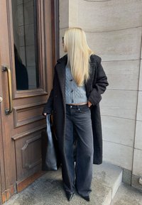 Zwarte oversized jas, grijze kabelgebreide trui, zwarte jeans met hoge taille, puntige zwarte hakken en een grote zwarte handtas.