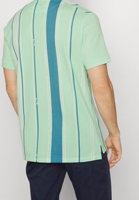 Polo shirt vert clair à manches courtes avec des rayures verticales bleues et blanches. Présente un petit logo près de l'ourlet et des fentes sur les côtés en bas.