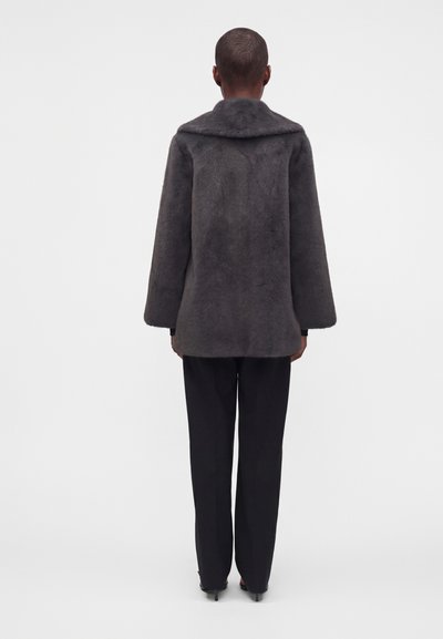 Personne debout, dos tourné, portant un manteau en fausse fourrure gris foncé, un pantalon noir et des chaussures noires à talons sur fond blanc.