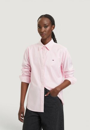 Femme aux cheveux tressés porte une chemise à rayures roses et blanches à boutons avec les manches retroussées et un pantalon noir, debout devant un fond uni.