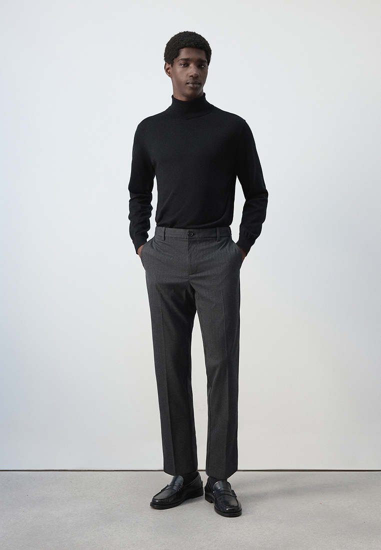 Jeune homme debout, les mains dans les poches, portant un col roulé noir, un pantalon gris et des mocassins noirs sur un fond uni.