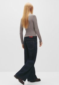 Grå struktureret langærmet top parret med løstsiddende mørke denim jeans. Outfittet har et rødt label på bagsiden af jeansene.