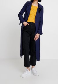 Námořnický trench coat s dvouřadým zapínáním, žluté tričko, černé zkrácené džíny a bílé tenisky. Hladká látka a klasický střih.