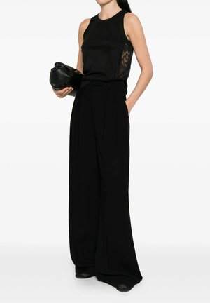 Donna che indossa un top nero senza maniche con inserti in pizzo ai lati, pantaloni neri a gamba larga, tiene in mano una pochette nera, una mano in tasca.