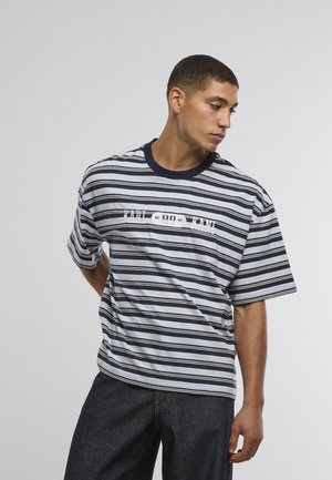 Muž oblečený v námornícky modrom a bielo pruhovanom tričku s krátkym rukávom a nápisom "KARL 89 KANI", v tmavých džínsoch, pozerajúci sa do strán na jednoduchom pozadí.