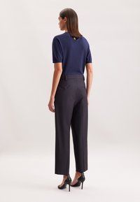 Vrouw die met haar rug naar de camera staat, gekleed in een marineblauw shirt met korte mouwen, zwarte hoge taille wijdlopende broek en zwarte hoge hakken.