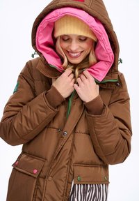 Brązowa puchowa kurtka z różowym podszewką, zielonym zamkiem i wielokolorowymi guzikami. Posiada kaptur oraz kieszenie z akcentami w wzór zebry.