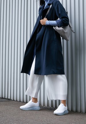 Personne portant un manteau bleu marine, un col roulé bleu clair, un pantalon blanc plissé, des baskets blanches, portant un sac à bandoulière gris en marchant devant un mur en métal ondulé.