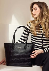 Sac fourre-tout noir en cuir crocodile gaufré avec deux poignées, présentant une forme rectangulaire et un branding subtil. Cardigan rayé partiellement visible.