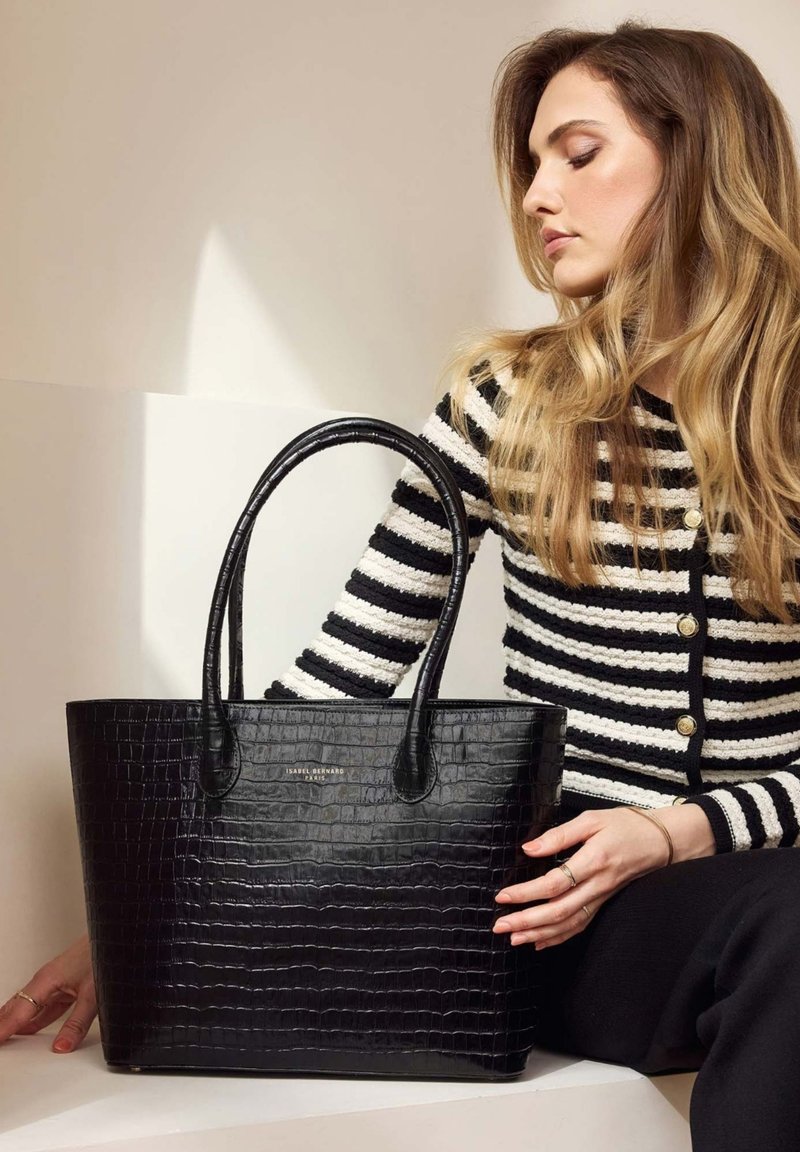 Sac fourre-tout noir en cuir crocodile gaufré avec deux poignées, présentant une forme rectangulaire et un branding subtil. Cardigan rayé partiellement visible.