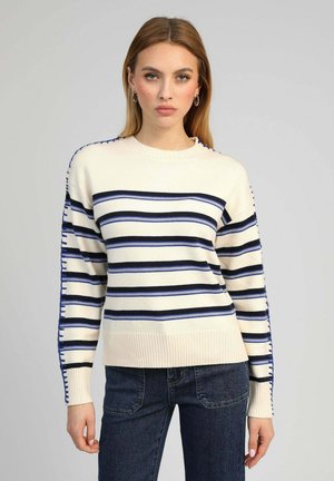 Femme portant un pull crème avec des rayures horizontales noires et bleues, associé à un jean bleu foncé, se tenant devant un fond uni.