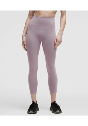Vrouw die een mauve cropped legging met hoge taille en zwarte sportschoenen draagt, staand tegen een effen lichtgrijze achtergrond.