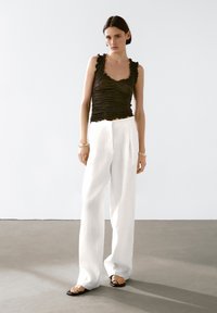 Black ruched tank top with ruffled straps, paired with high-waisted white linen trousers. Model wears flat black sandals. Minimalist background.