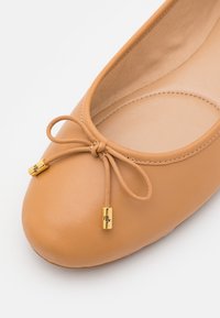 Ballerine in pelle color cuoio con punta rotonda, caratterizzata da un dettaglio a fiocco e accenti in metallo dorato alle estremità del fiocco.