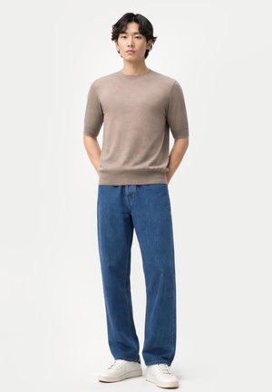 Junger Mann steht mit Blick nach vorne, trägt einen beigen Kurzarmpullover, blaue Jeans und weiße Sneakers vor einem schlichten Hintergrund.