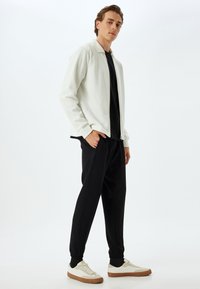 Weiße Zip-Jacke mit geripptem Kragen, schwarzes T-Shirt darunter, schwarze schmal geschnittene Jogginghosen und beiges Sneakers mit Gummisohlen.