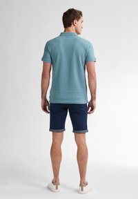 Camisa polo azul de mangas cortas, combinada con pantalones cortos azul marino con dobladillos enrollados. Zapatillas blancas con acentos beige. Vista trasera.