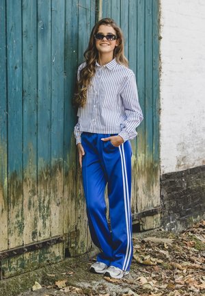 Blauwe trainingsbroek met witte strepen, een wit gestreept overhemd met knopen, en grijze sneakers, staand voor een verweerde houten achtergrond.