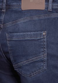 Dark blue denim jeans feature a back pocket with a metal button and contrasting stitching. Leather brand patch on waistband.