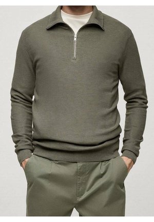 Hombre con un suéter con cremallera texturizado de color verde oliva sobre una camisa blanca, con pantalones a juego verde oliva, con las manos en los bolsillos.