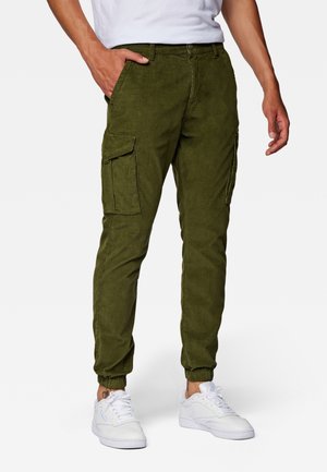 Man die olijfgroene cargobroek met elastische boorden en witte sneakers draagt, staat met één hand in de zak tegen een effen achtergrond.