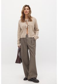 Cardigan lavorato a maglia beige con cintura frontale, abbinato a pantaloni grigi larghi. Il modello tiene una borsa bordeaux e indossa mocassini neri.