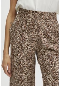 Kobieta w spodniach o szerokiej nogawce w panterkę z zebranym elastycznym pasem, stojąca z lewą ręką swobodnie opuszczoną wzdłuż ciała.