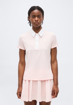 Noor naine lühikeste loomulike juustega, kes kannab kahvaturoosat lühikeste varrukatega polosärki ja sobivat volditud seelikut, seisab ühevärvilise tausta ees.