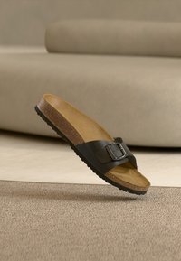 Sandale à glissière en cuir noir avec une seule boucle ajustable, semelle en liège et semelle en caoutchouc texturée, suspendue au-dessus d'un fond neutre.