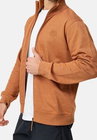 Veste zippée en orange terreux avec une texture côtelée, col montant et logo discret. Elle est fabriquée en tissu tricoté avec des poignets élastiques, portée par-dessus une chemise blanche.
