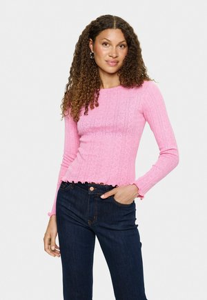 Vrouw met krullend haar, gekleed in een roze lange mouwen gebreide top en donkerblauwe jeans, met één hand in haar zak tegen een witte achtergrond.