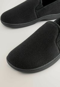 Juodos slip-on bateliai su tekstūruotu audiniu herringbone ir elastingais šonais. Guminė padas suteikia sukibimą ir komfortą.