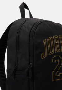 Mochila negra con dos compartimentos con cremallera, bordado "JORDAN 23" en dorado, fabricada con material duradero y con asa de transporte en la parte superior.