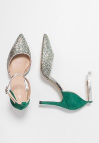 Gröna mockaklackar med silverglittriga detaljer och ankelband, uppvisade mot en vit bakgrund.