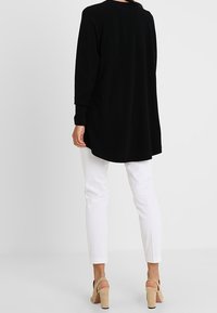 Svart långärmad tunika med rundad halsringning och sidofickor, tillsammans med vita åtsittande byxor och beige klacksandaler.